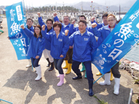 港で青いジャンパーを着たメンバーが魚の日の旗を持ってポーズをとる写真