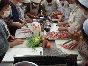 三角巾とエプロン姿の複数の人が調理台を囲み、野菜を包丁で切っている写真
