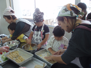 三角巾とエプロン姿の大人と子どもが並び、調理台で協力して料理を作っている写真