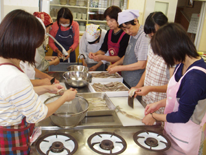 エプロン姿の複数の人が調理台を囲み、包丁で食材を切り分担して調理している写真