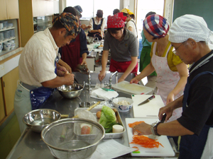 三角巾とエプロン姿の複数の人が調理台を囲み、野菜を包丁で切り分担して料理している写真
