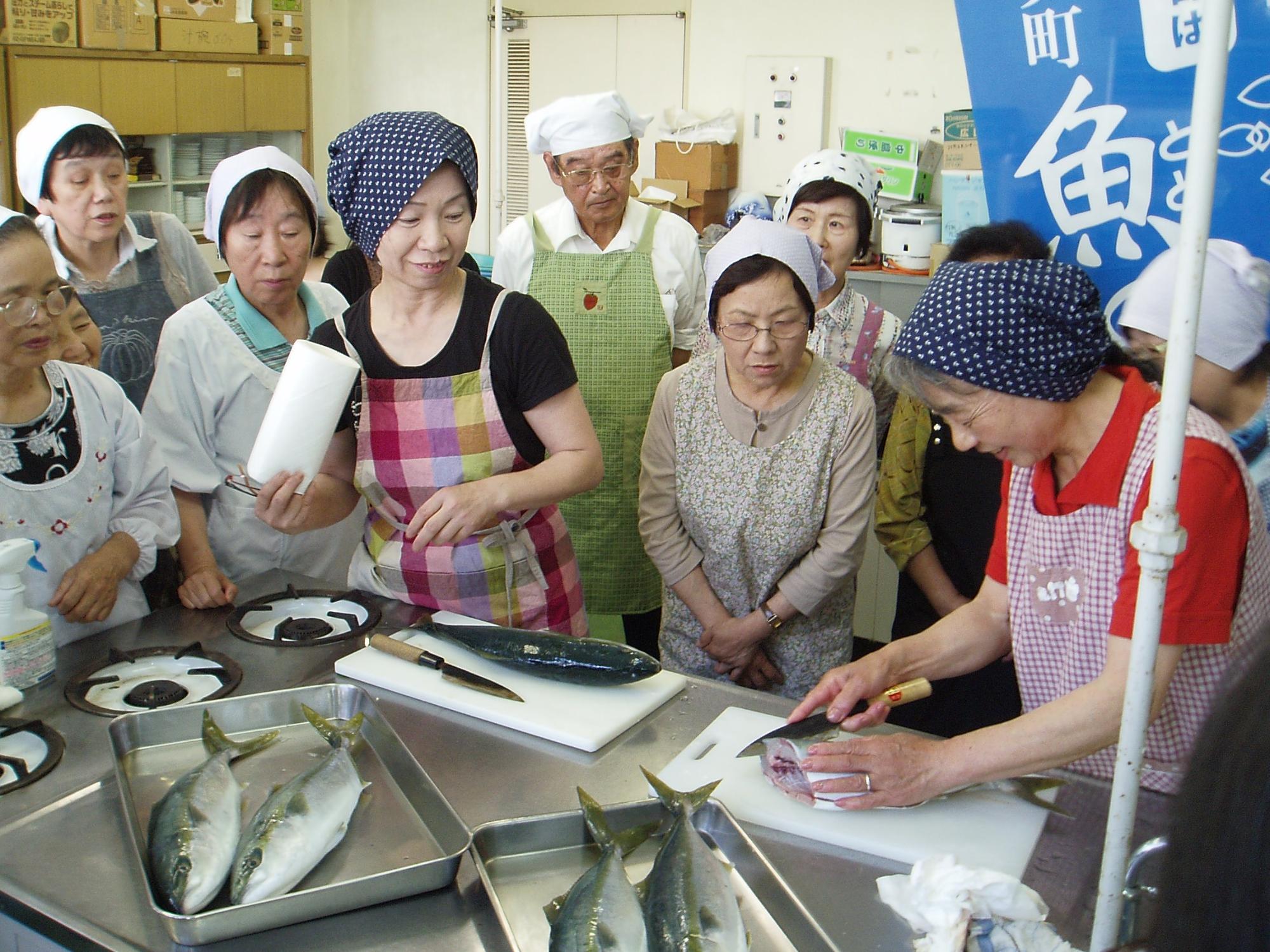 調理室で参加者が集まり魚のさばき方の説明を聞いている写真