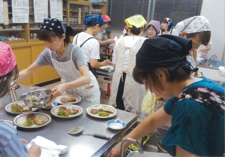 調理室でエプロンと三角巾を着けた複数の人が料理を盛り付けている写真