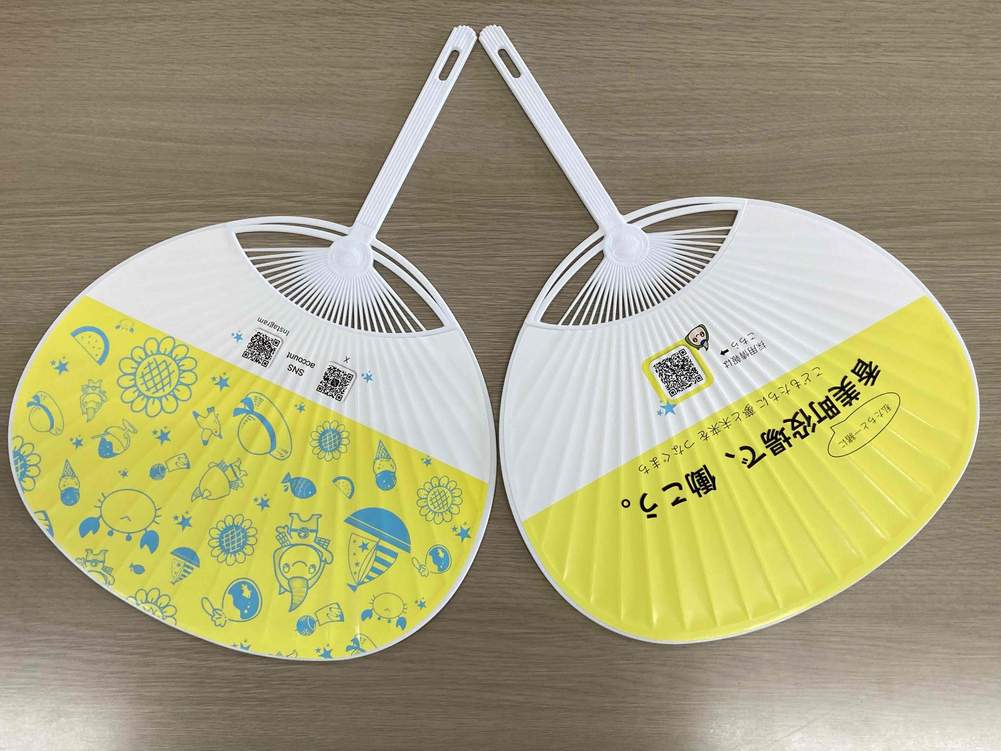 黄色の下地に青色で描かれた様々なイラストがあり、InstagramやXのQRコードがついている面と、黄色の下地に「香美町役場で、働こう」と黒字で書かれ採用情報へのQRコードが記された面が並んでいる職員募集PRうちわの写真