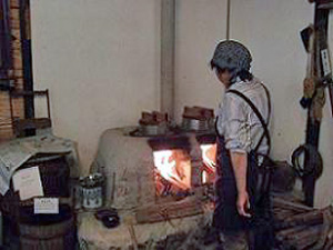 かまどの番をするエプロン姿の人の後ろ姿の写真