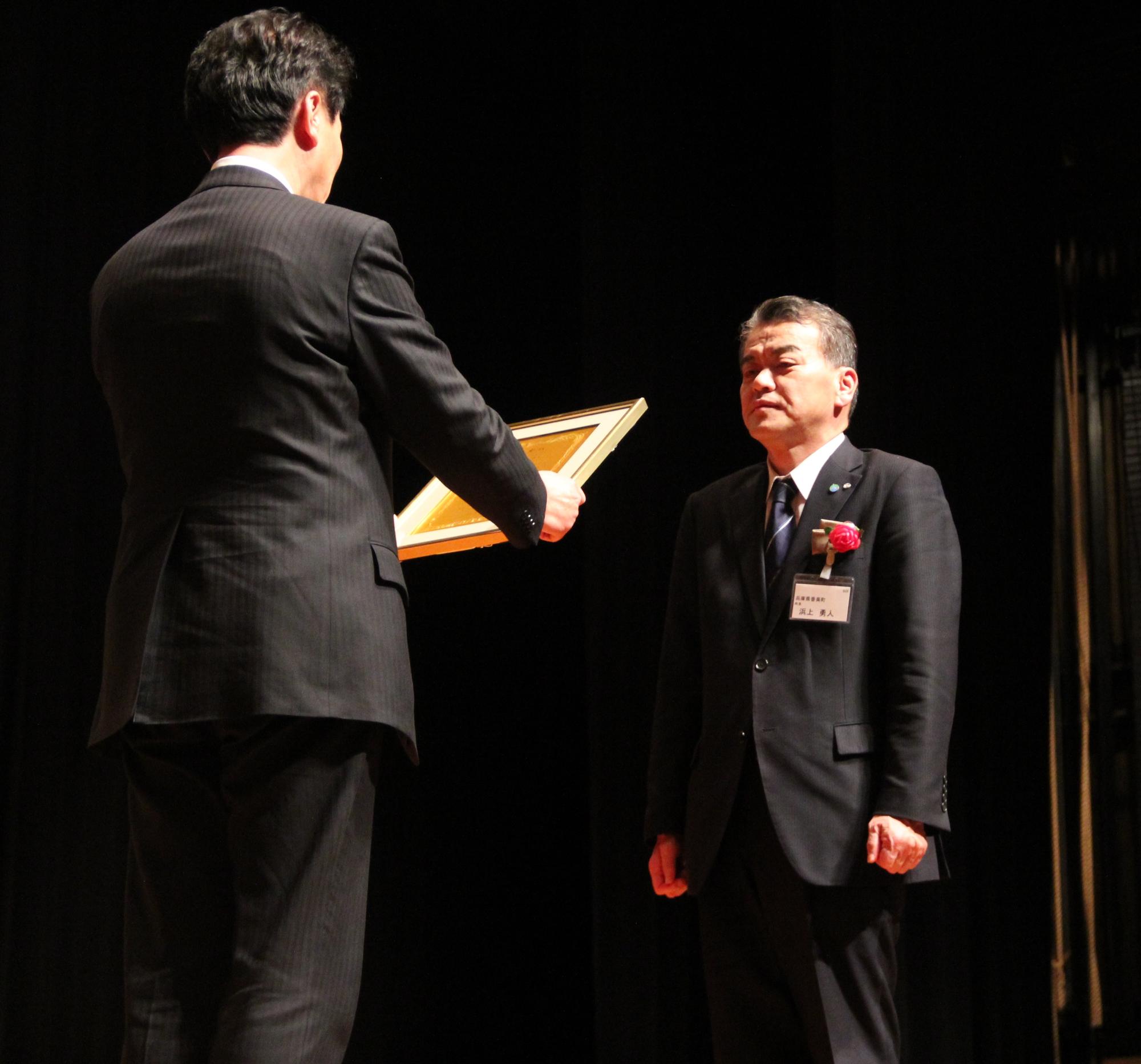 額縁に入った賞状を手に持つスーツ姿の男性と向かい合って立つ胸元に赤い造花をつけたスーツ姿の男性の写真