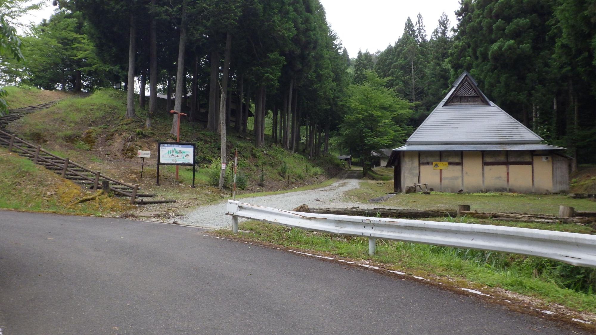 道路の奥の森の入り口に建つ1件の建物の写真