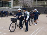 校庭で体操服の生徒たちが自転車に乗りながら交通安全指導を受けている様子の写真