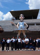 大きな建物の前に建てられた人形のモニュメントの前で中学生たちが並んで集合写真を撮っている写真