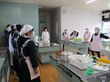 家庭科室で調理実習の説明を聞くエプロン姿の生徒たちの写真