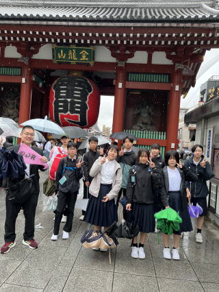 雨がかすかに降る浅草雷門前で、ポーズを取っている制服姿の学生たちと引率の先生が集合写真を撮っている様子の写真