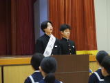 体育館の壇上でたすきを掛けた男子生徒二人が演台からあいさつしている写真