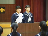 体育館の壇上でたすきを掛けた女子生徒二人が演台からあいさつしている写真