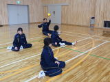 体育館の床に座った生徒たちがボール投げの練習をしている写真