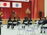 体育館の舞台上で校長先生が国旗と校旗を背に式辞を述べ生徒たちが着席して聞いている写真