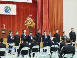 体育館の舞台前に制服姿の生徒たちが一列に並び来賓席の前で式典に臨んでいる写真