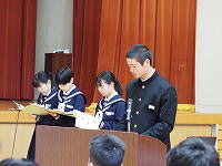 体育館の壇上で制服姿の生徒が原稿を読み上げて発表している様子の写真