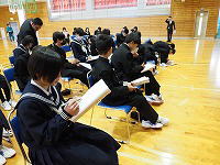 体育館で制服姿の生徒たちが椅子に座って資料を読みながら説明を聞いている様子の写真