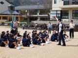 運動場で体操服の生徒たちが地面に座って説明を聞いている様子の写真