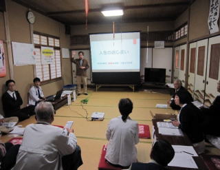 和室の前方のスクリーン横に茶色の服を着た男性が手を挙げて立って話している写真