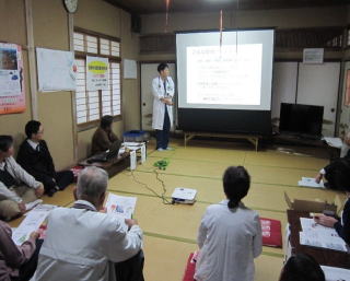 和室の前方のスクリーン横に白衣を着た男性が立って話し、地域の方が聞いている様子の写真