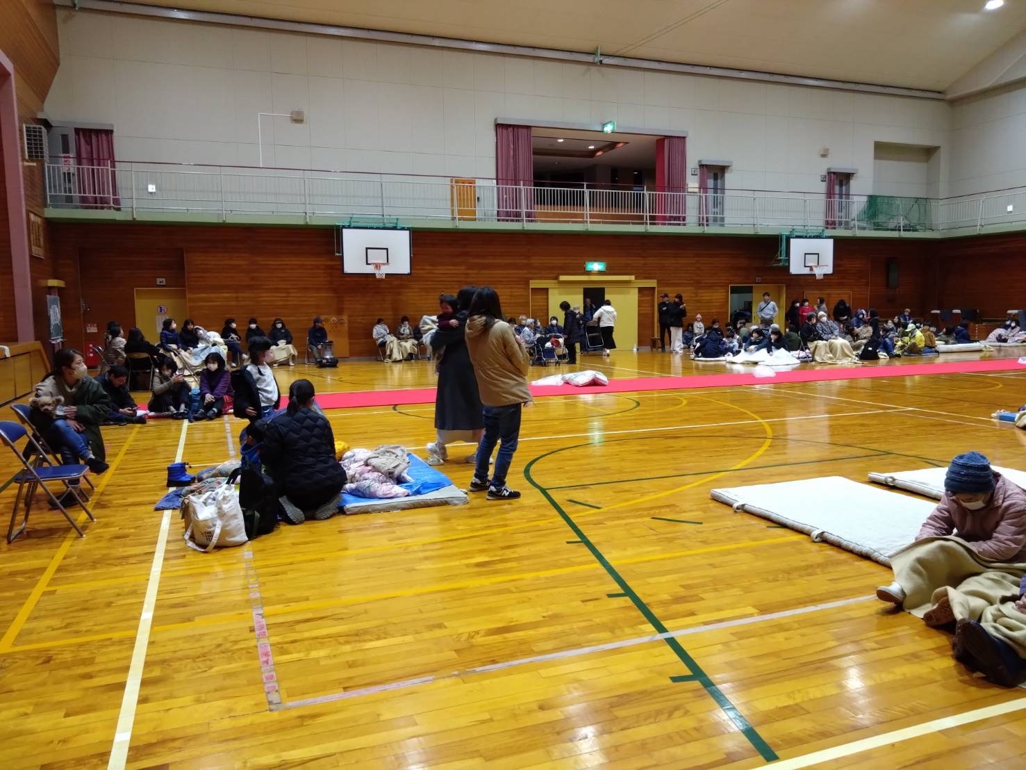 小学校の体育館でマットや毛布を敷いて身を寄せ合う避難者たちの写真
