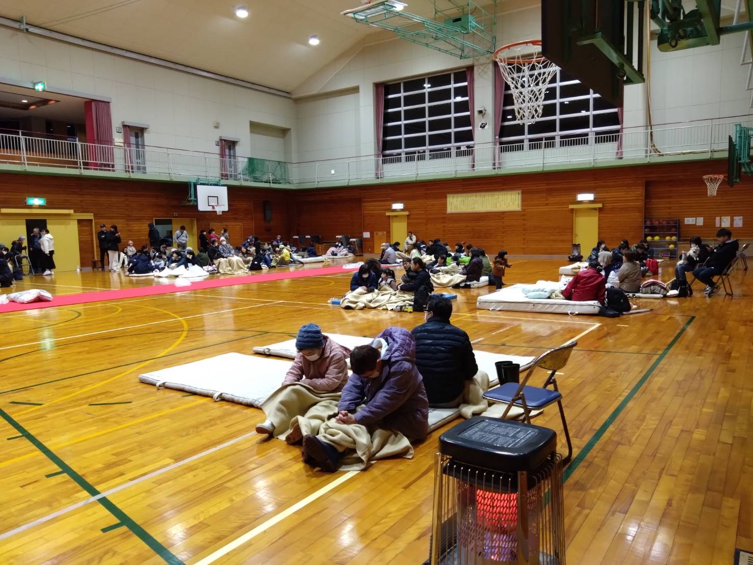 避難所の体育館で、ダウンジャケットなどの防寒着を着た人々が、床に座っている写真