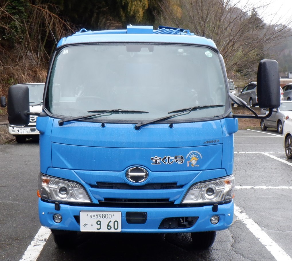 背景に山がある駐車場に、宝くじ号のロゴが入った青いもみ収集車が正面から写されている写真