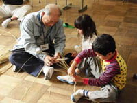 室内で男性が児童2人にしめ縄づくりを教えている様子の写真