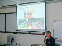 室内でマイクを持った男性がスクリーンを用いて説明を行っている様子の写真