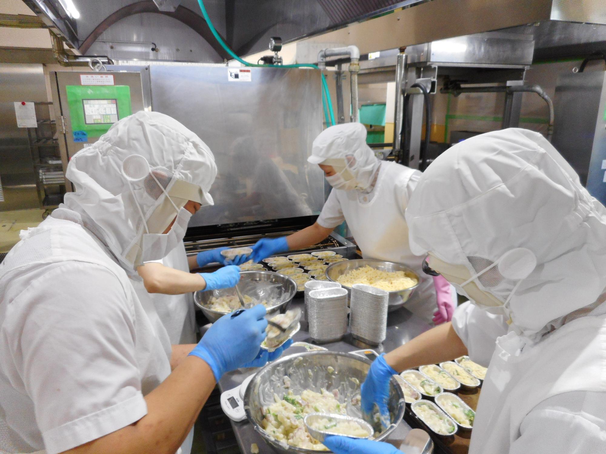 室内で調理員たちがボウルを用いて具材を配置している様子の写真