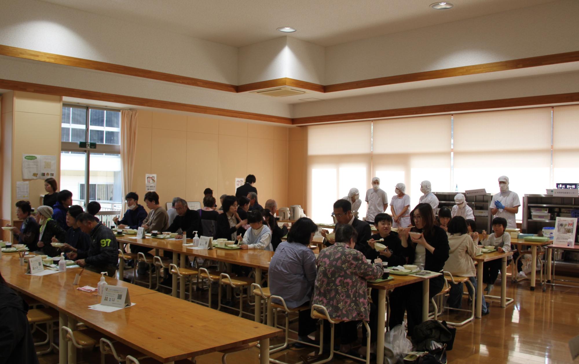 室内で4つに並んだ机の上で大人たちが食事を取っている様子の写真