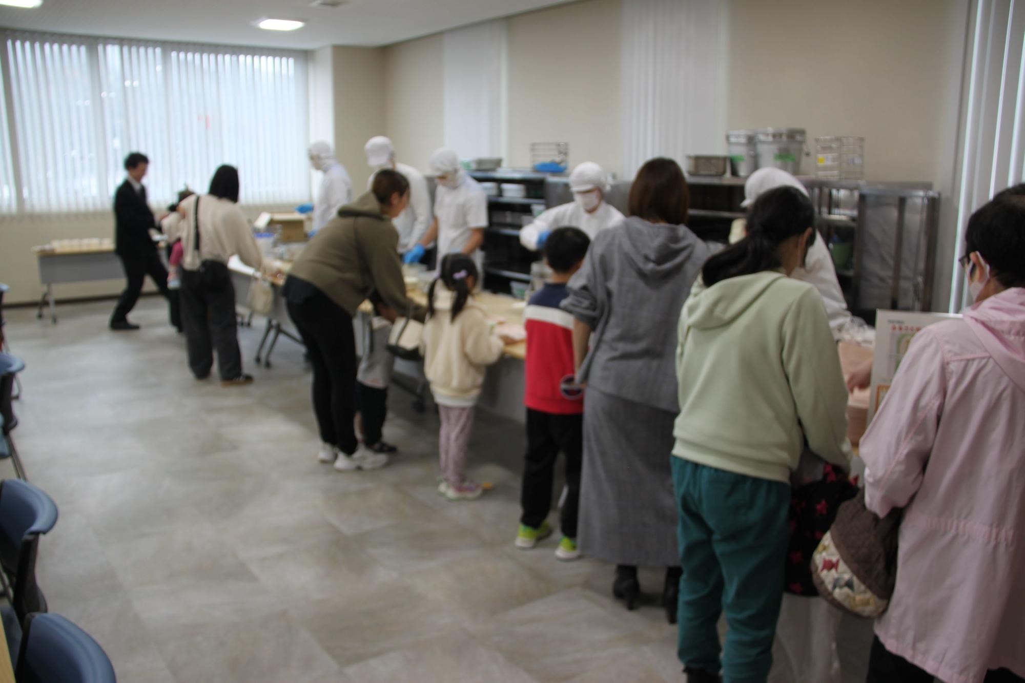 割烹着を着た人たちが並んでいる親子たちに給食を配膳している写真