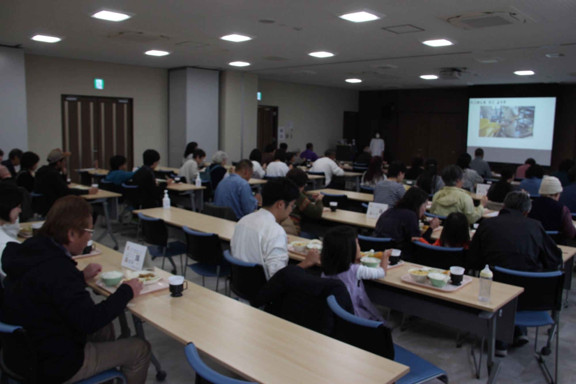 手前にスクリーンがある部屋で長机で給食を食べている親子たちの写真