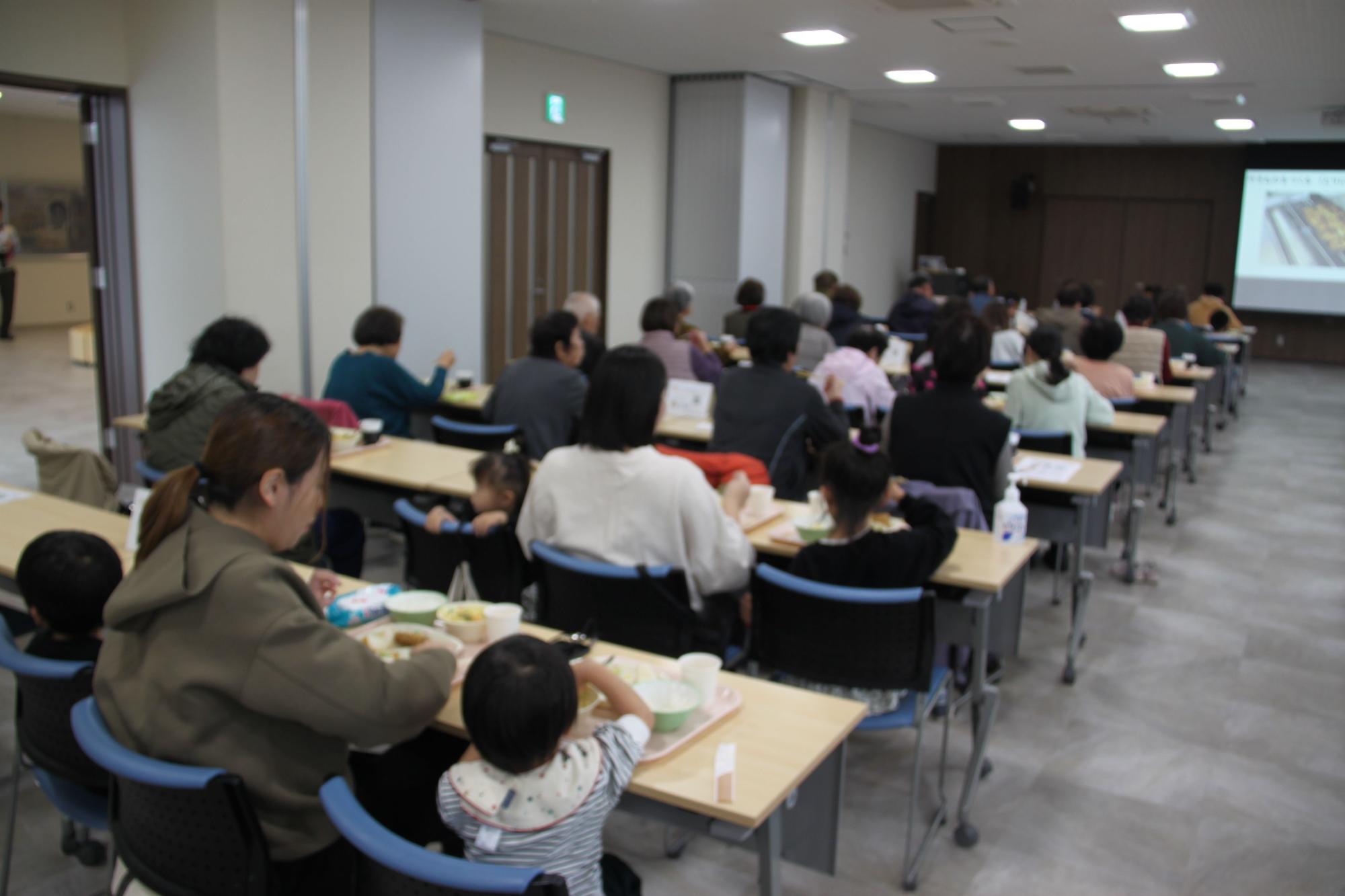 手前にスクリーンがある部屋で長机で給食を食べている母親と小さな娘さんの写真