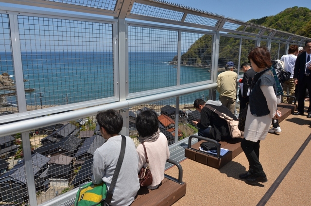 高台のベンチに座って港町を眺める人たちの写真