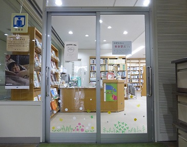 ガラス扉越しにカウンターや本棚が並ぶ図書コーナーの室内が見えている写真