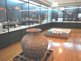 部屋の中央に大きな土器があり、部屋の壁沿いに設置されたショーケースに様々な土器などが展示されている写真