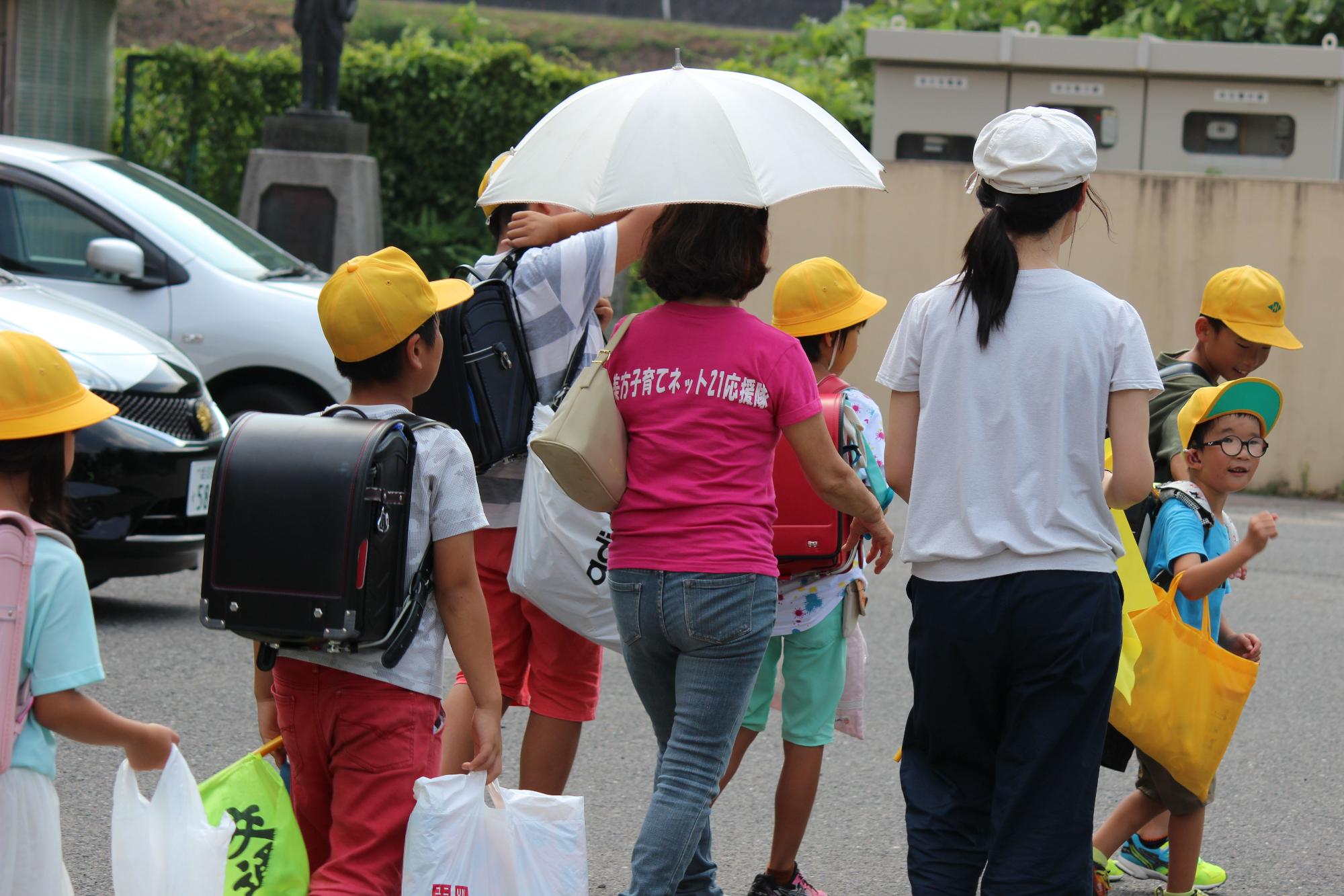 黄色の帽子を被った児童たちの傍を歩く二人の女性の写真