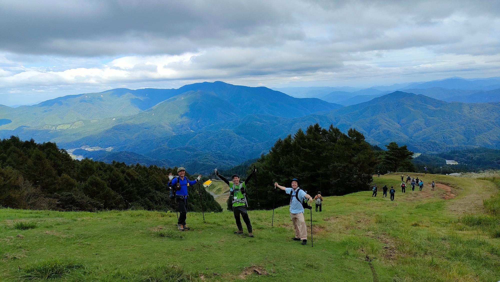 高い場所にある草原で両手を広げて立っている3人の登山客の写真