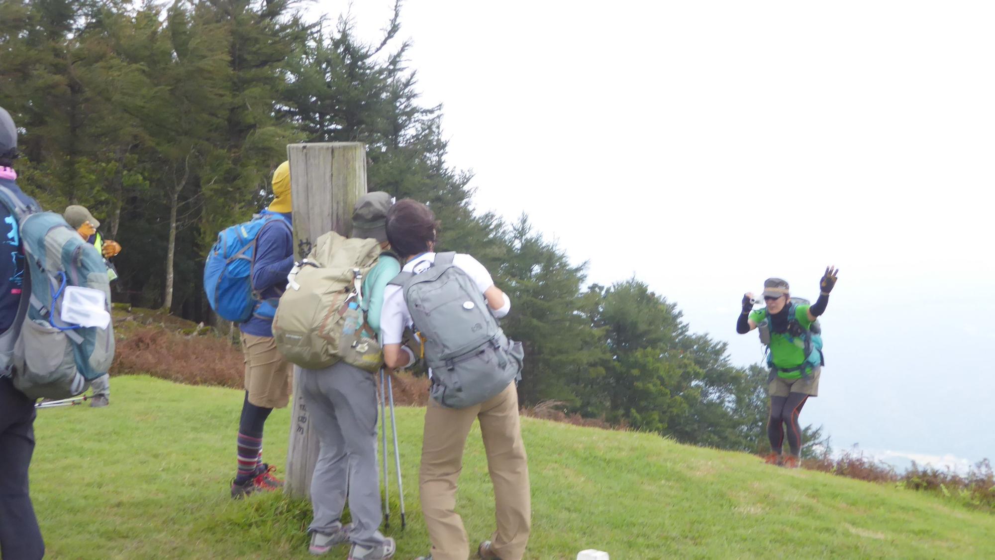 山頂にある標識に集まっている3人を撮影する男性の写真