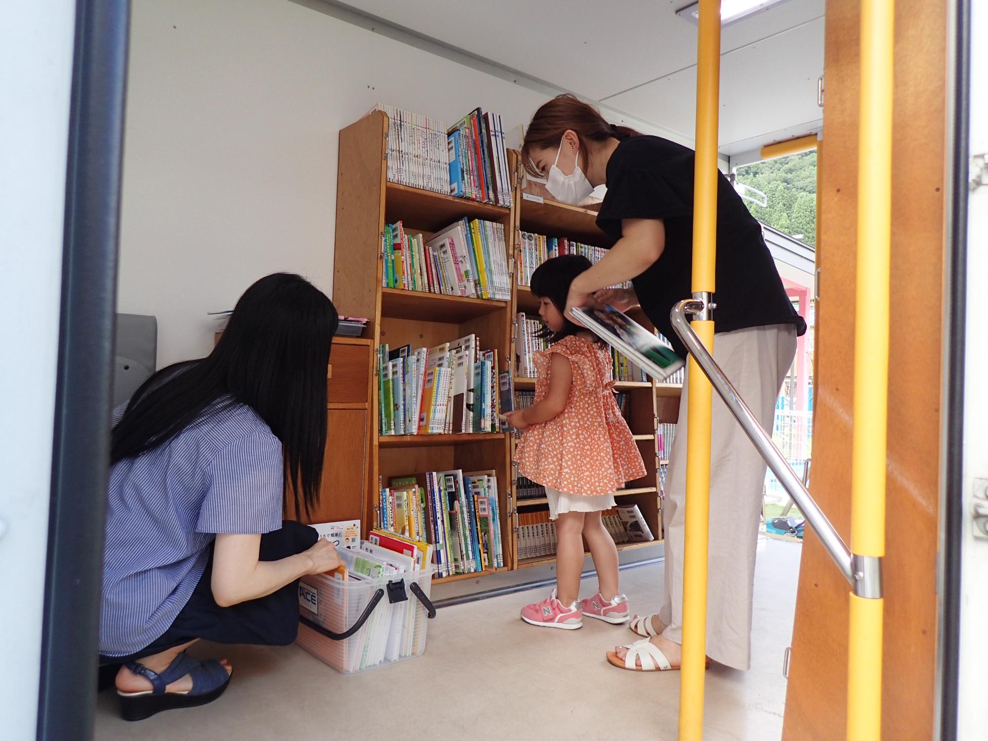 箱の中の本を選ぶ女性を見ている子供と女性の写真