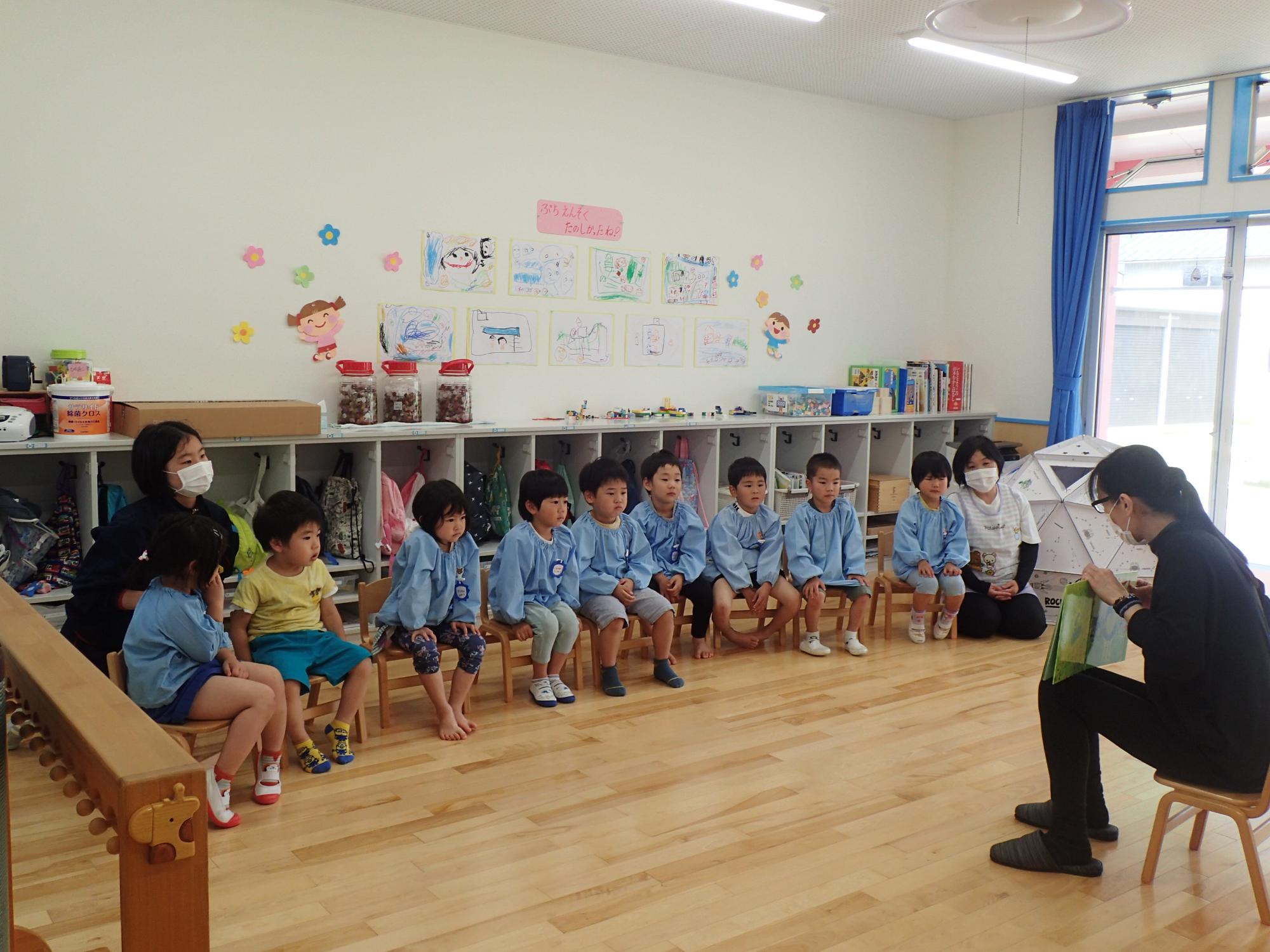 スモックを着た園児たちが絵本の読み聞かせを聞いている写真