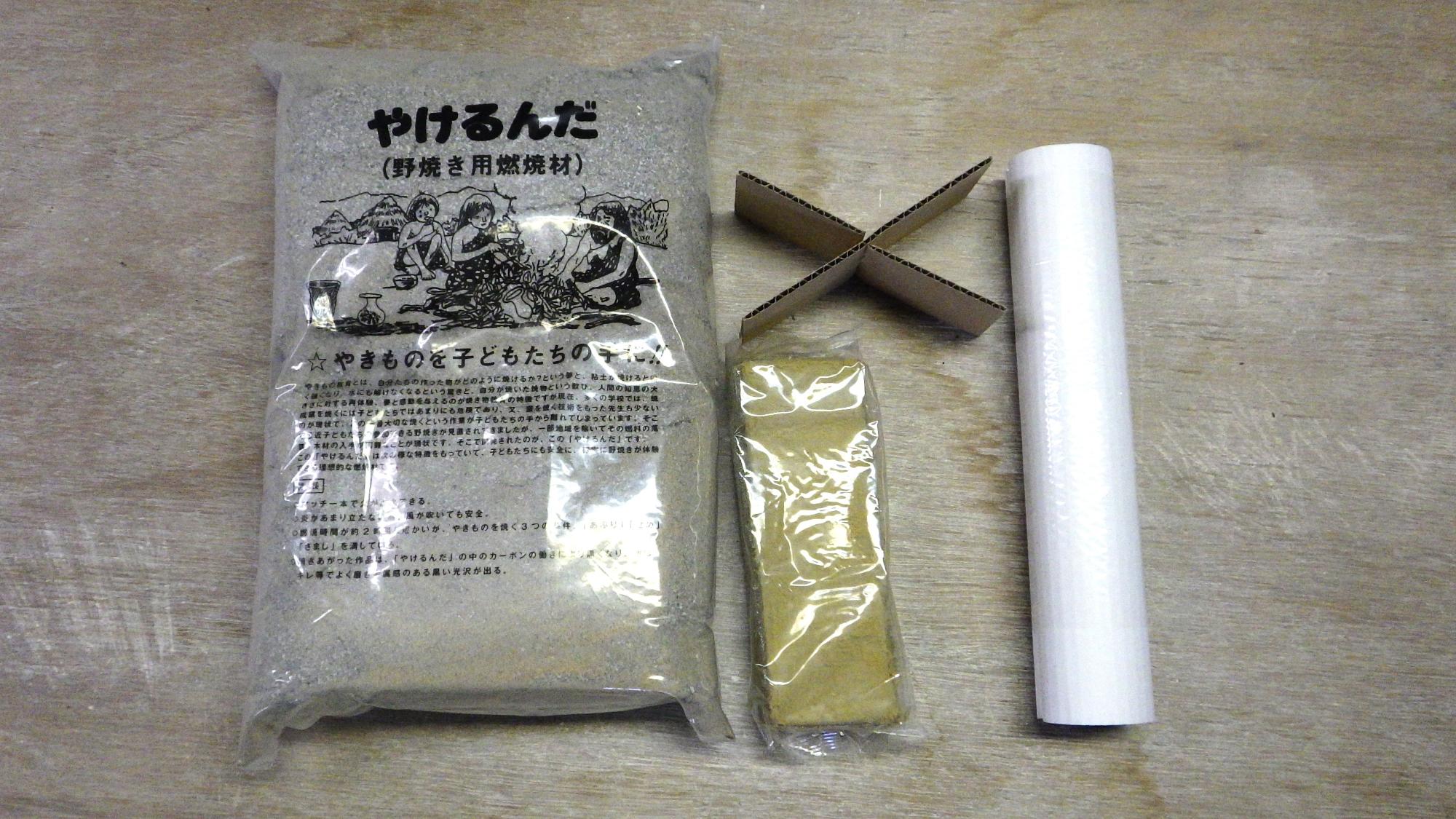 黄土色の粘土と「やけるんだ」と書かれた野焼き用燃焼材と十字に組まれた段ボールと白い筒の写真