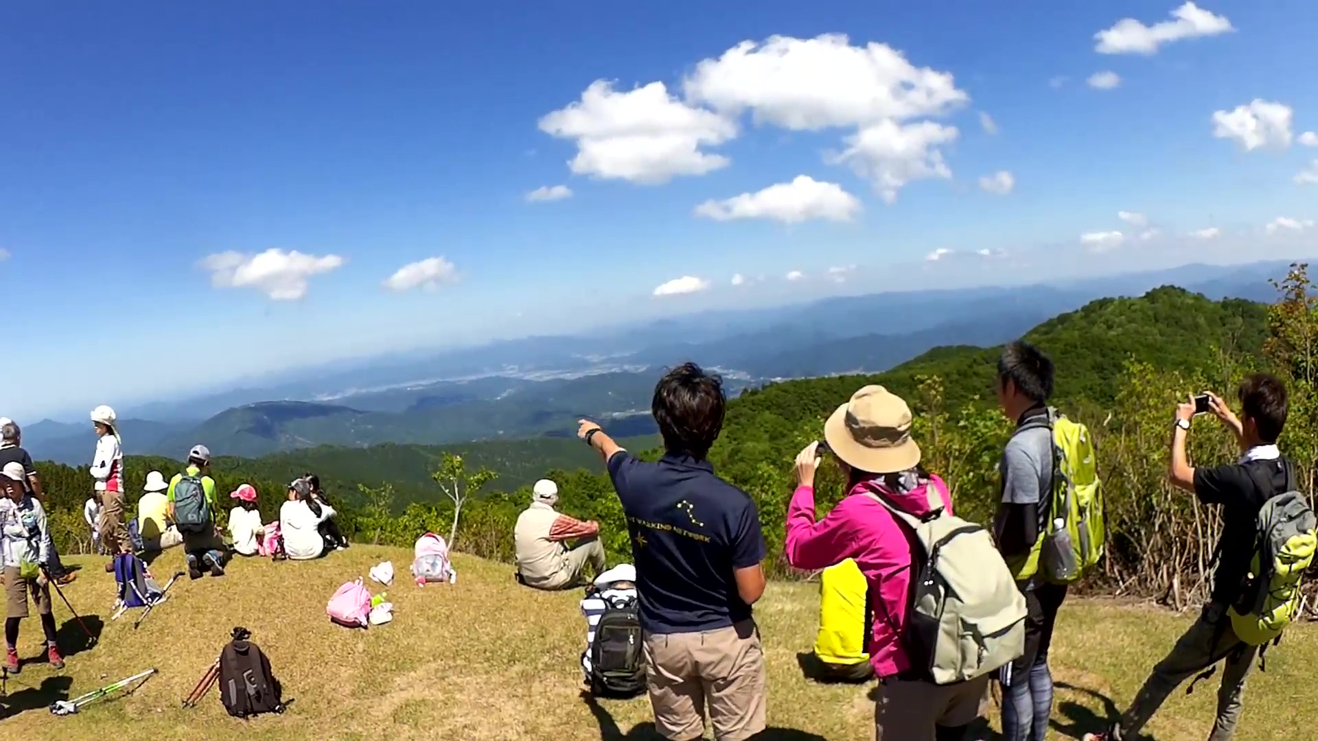 山の開けたところで遠くの山を指さす登山客の写真