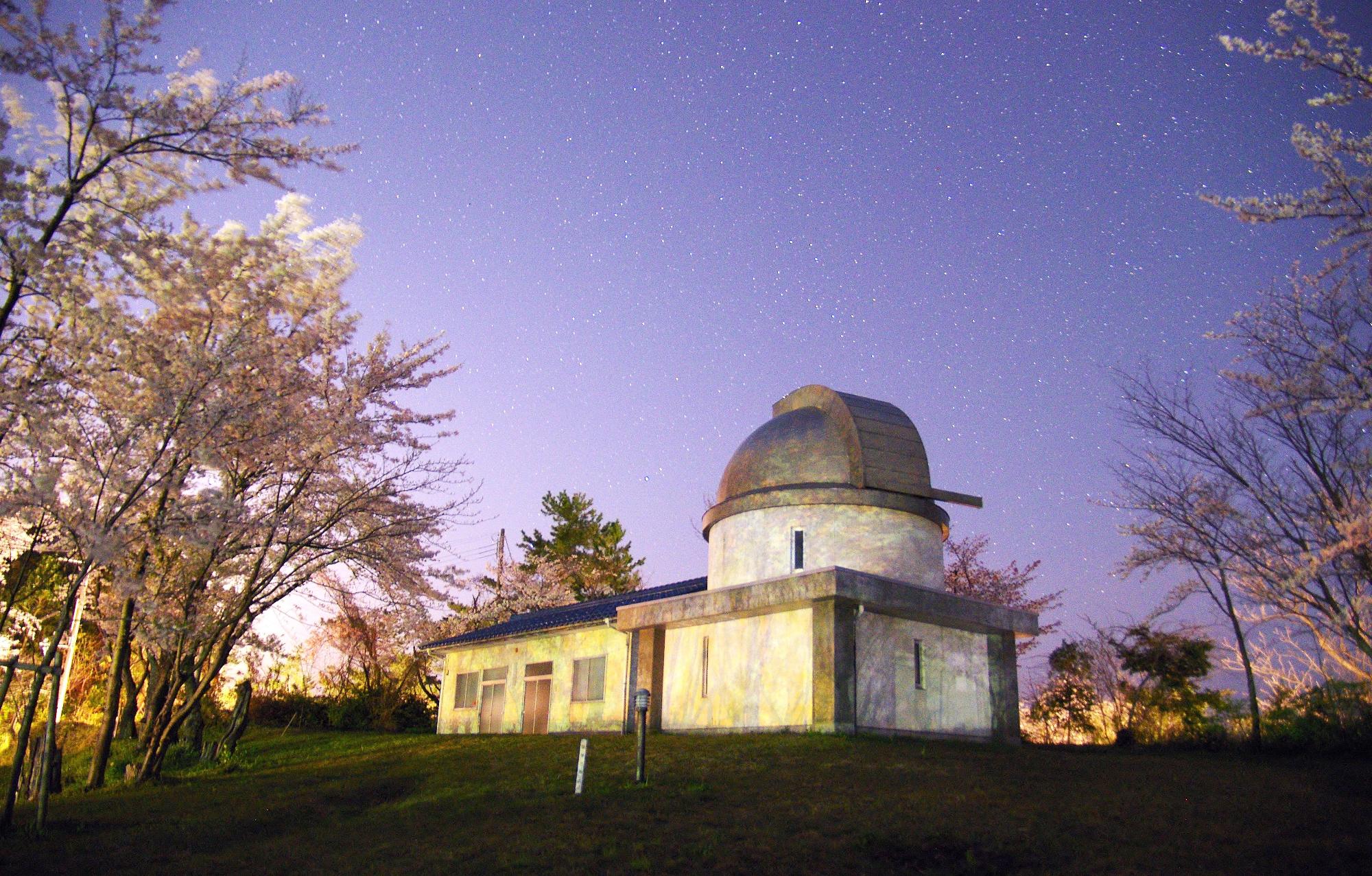 満天の星空の下、桜の木に囲まれたドーム屋根の天文台の写真
