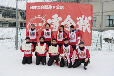 雪合戦と書かれた赤旗の前で賞状やトロフィーを見せて並ぶ人々の集合写真