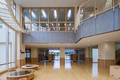 吹き抜けの広いホールに木製フロアと丸テーブルが配置された校舎内ロビーの写真