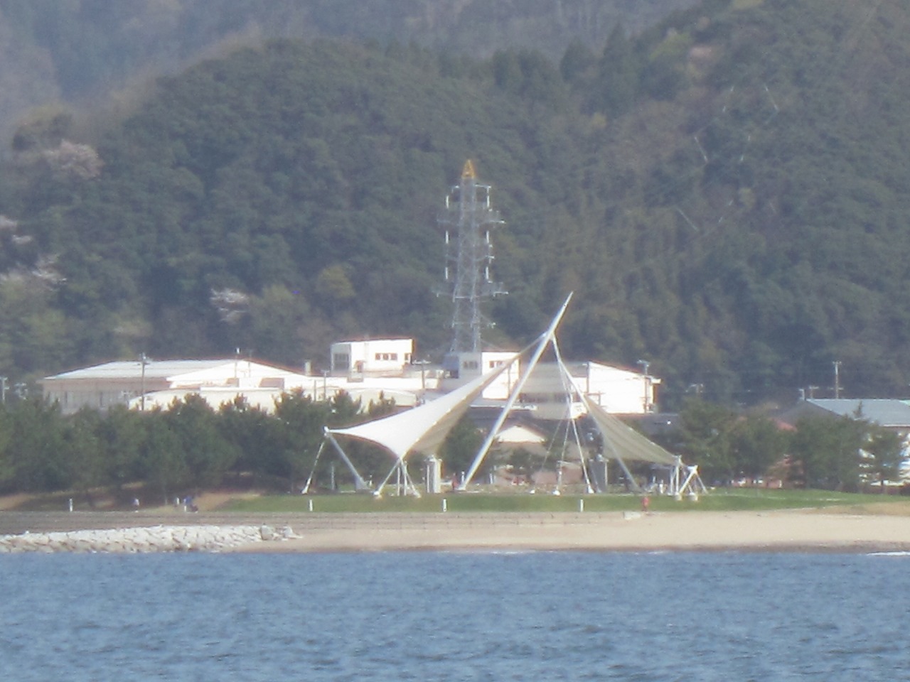 沿岸部にある周りを山に囲まれた芝生の広場に白いシャフトと布で表現されたモニュメントが設置されている写真