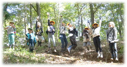 光がキラキラと差し込む明るい雑木林の中で、10人ほどの子どもたちが横一列に並んで、地面を力いっぱい蹴り上げ、空に向かって大ジャンプをしている瞬間を捉えた写真